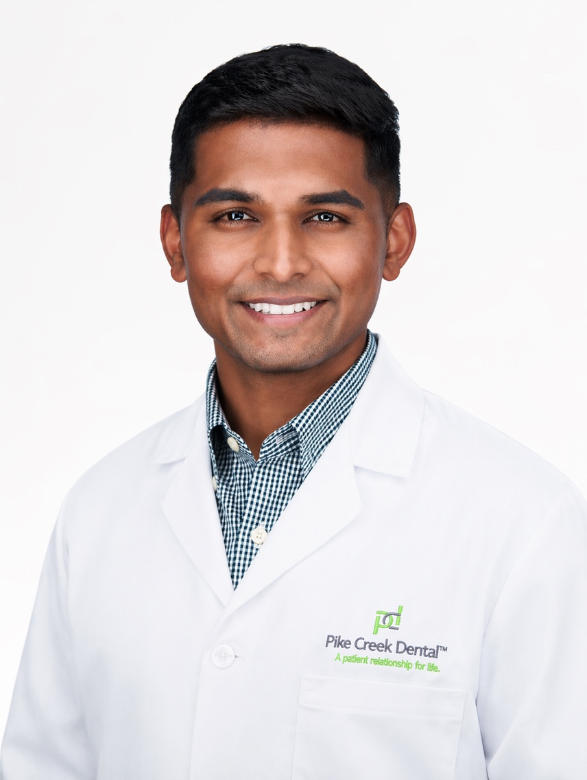 Smiling dentist in a white coat, Pike Creek Dental logo visible on the chest.